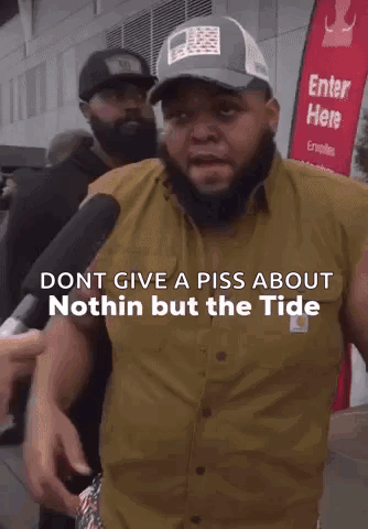 A Man With A Beard Is Talking Into A Microphone While Another Man Stands Behind Him .. GIF