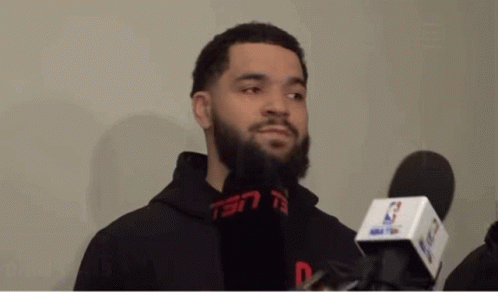 A Man With A Beard Is Talking Into A Microphone While Wearing A Black Hoodie . GIF