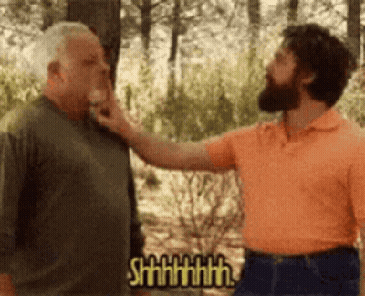 A Man With A Beard Is Touching The Face Of Another Man In A Forest . GIF