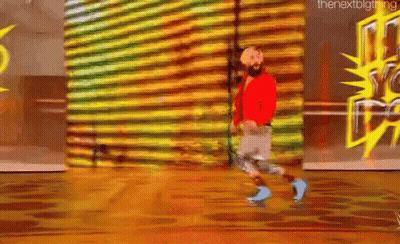 A Man With A Beard Is Walking On A Wooden Floor In Front Of A Wall With Graffiti On It . GIF