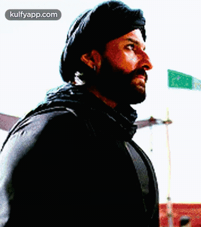 A Man With A Beard Is Wearing A Black Hat And Scarf And Looking To The Side . GIF