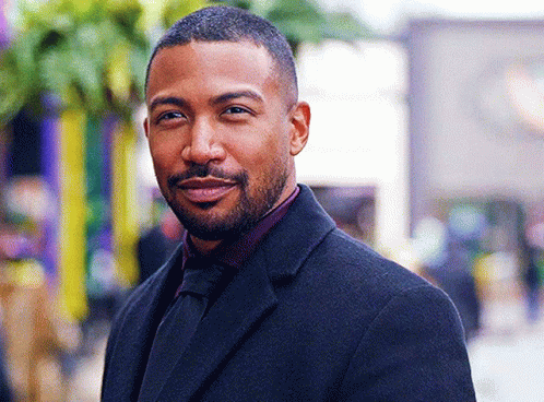 A Man With A Beard Is Wearing A Black Suit And Tie And Smiling . GIF