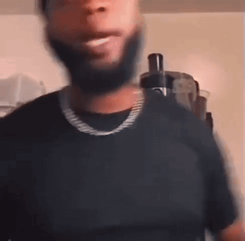 A Man With A Beard Is Wearing A Black T-Shirt And A Gold Chain Around His Neck . GIF