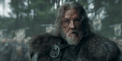 A Man With A Beard Is Wearing A Fur Coat And A Badge With A Cross On It . GIF