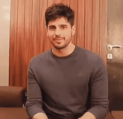 A Man With A Beard Is Wearing A Grey Sweater And Smiling While Sitting On A Couch . GIF