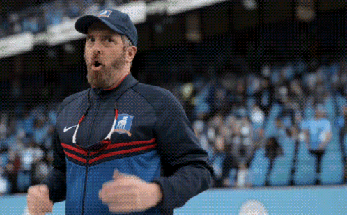 A Man With A Beard Is Wearing A Hat And A Jacket While Standing In Front Of A Crowd . GIF