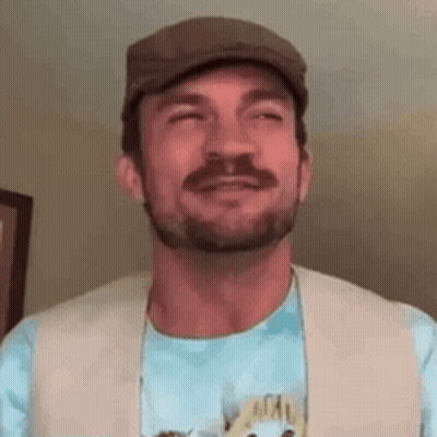 A Man With A Beard Is Wearing A Hat And A Vest And Smiling . GIF
