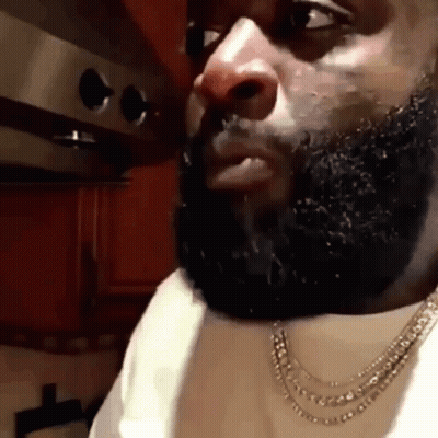A Man With A Beard Is Wearing A Necklace And A White Shirt In A Kitchen . GIF