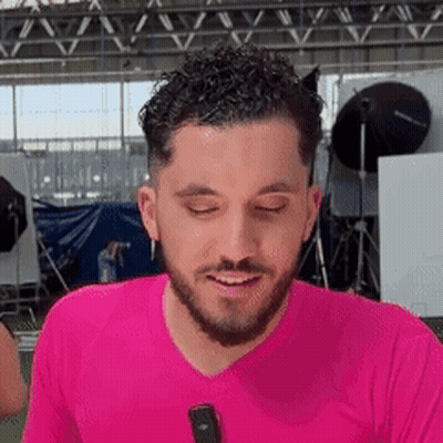 A Man With A Beard Is Wearing A Pink Shirt And Smiling While Holding A Microphone . GIF