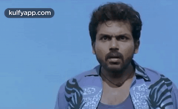 A Man With A Beard Is Wearing A Purple Shirt And Looking At The Camera With A Blue Sky In The Background . GIF