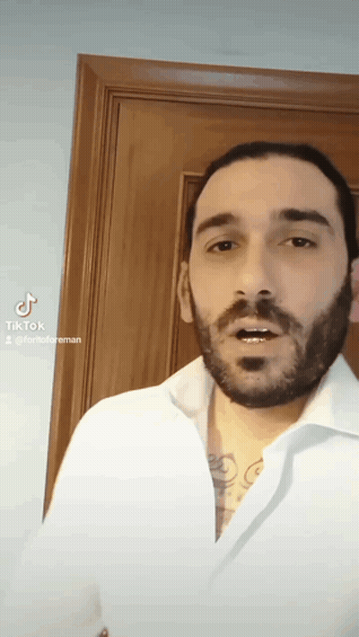 A Man With A Beard Is Wearing A White Shirt And A Tattoo On His Chest . GIF