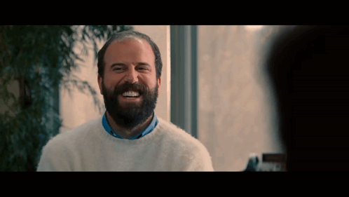 A Man With A Beard Is Wearing A White Sweater And Smiling GIF