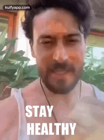 A Man With A Beard Is Wearing A White Tank Top And A Necklace . GIF
