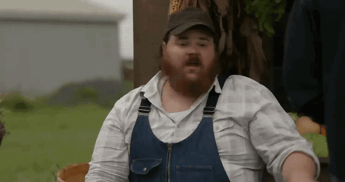 A Man With A Beard Is Wearing Overalls And A Baseball Cap . GIF