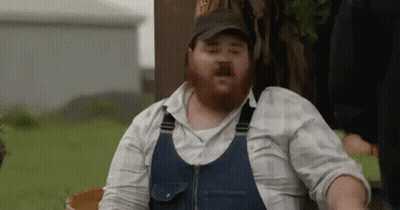 A Man With A Beard Is Wearing Overalls And A Hat While Sitting In A Field . GIF