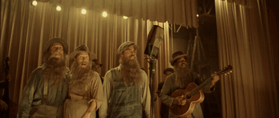 A Man With A Beard Sings Into A Microphone While Other Men Play Instruments GIF
