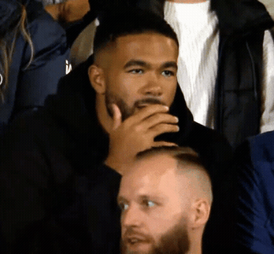A Man With A Beard Sits In A Crowd With His Hand To His Mouth GIF