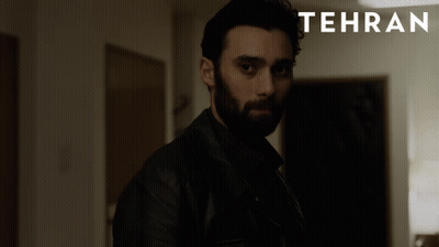 A Man With A Beard Stands In A Dark Room With Tehran Written On The Bottom GIF