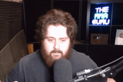 A Man With A Beard Stands In Front Of A Microphone In Front Of A Sign That Says The Mma Guru GIF
