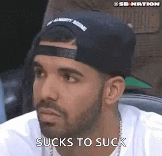 A Man With A Beard Wearing A Baseball Cap And A White Shirt Is Sitting In A Chair . GIF