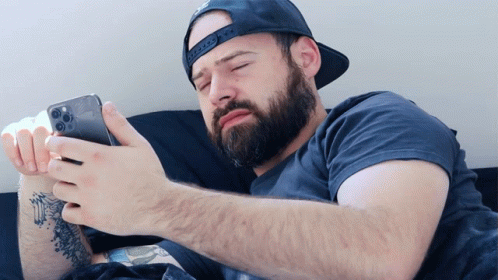A Man With A Beard Wearing A Baseball Cap Looks At His Phone GIF