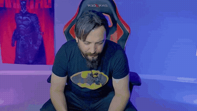 A Man With A Beard Wearing A Batman Shirt Is Sitting In A Chair GIF