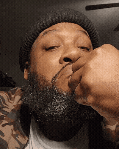 A Man With A Beard Wearing A Beanie Is Holding His Fist To His Face GIF