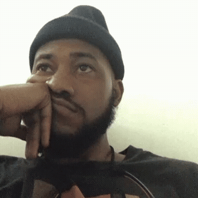 A Man With A Beard Wearing A Black Beanie Is Sitting Down With His Hand On His Face . GIF