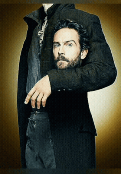A Man With A Beard Wearing A Black Coat Has His Hand On His Pocket GIF