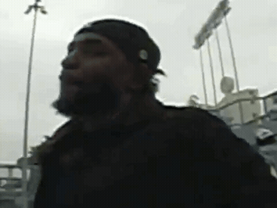 A Man With A Beard Wearing A Black Hat Is Standing In Front Of A Baseball Stadium . GIF