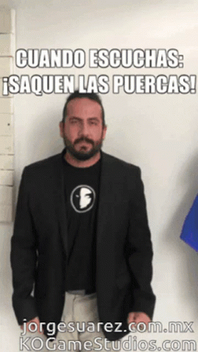 A Man With A Beard Wearing A Black Jacket And A Black Shirt With The Letter F On It Stands In Front Of A White Wall GIF