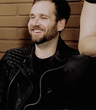 A Man With A Beard Wearing A Black Leather Jacket Is Smiling While Sitting In Front Of A Brick Wall . GIF
