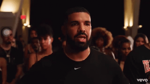 A Man With A Beard Wearing A Black Nike Shirt Stands In Front Of A Crowd Of People . GIF