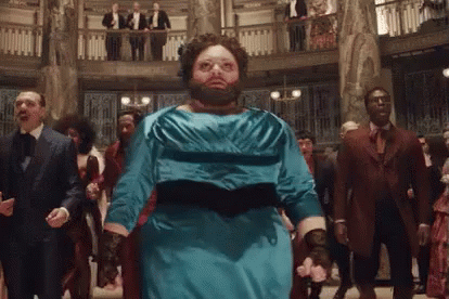 A Man With A Beard Wearing A Blue Dress Is Dancing In A Room With Other People . GIF