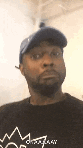 A Man With A Beard Wearing A Blue Hat And A Black Shirt With A Crown On It . GIF