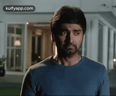 A Man With A Beard Wearing A Blue Shirt Is Standing In Front Of A Building . GIF