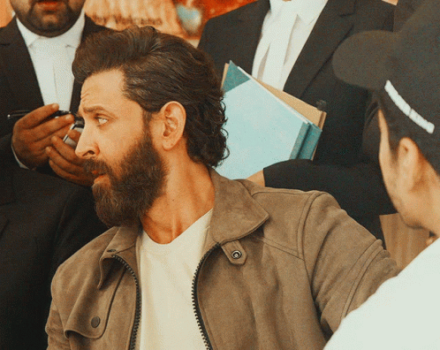 A Man With A Beard Wearing A Brown Jacket Is Talking To Another Man GIF