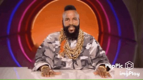 A Man With A Beard Wearing A Camouflage Jacket And Gold Chains Is Sitting At A Table With His Hands On The Table GIF