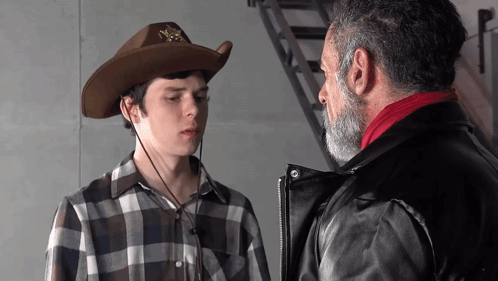 A Man With A Beard Wearing A Cowboy Hat Talks To Another Man In A Plaid Shirt GIF