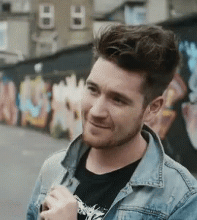 A Man With A Beard Wearing A Denim Jacket And A Black Shirt Is Smiling In Front Of A Graffiti Wall . GIF