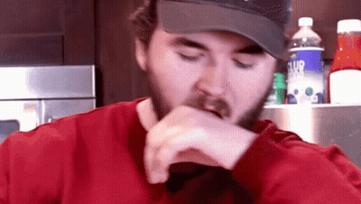 A Man With A Beard Wearing A Hat And A Red Shirt Eating GIF