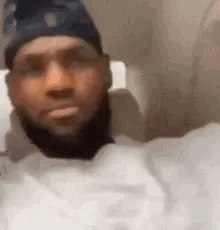 A Man With A Beard Wearing A Hat And A White Shirt Is Sitting In A Chair . GIF