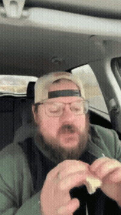 A Man With A Beard Wearing A Hat And Glasses Is Eating A Sandwich GIF
