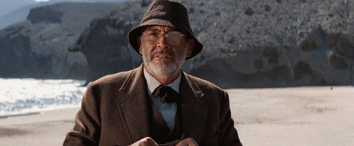 A Man With A Beard Wearing A Hat And Glasses Stands On A Beach GIF