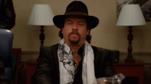 A Man With A Beard Wearing A Hat And Scarf Is Sitting At A Table GIF