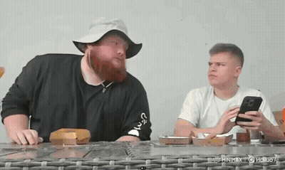 A Man With A Beard Wearing A Hat Is Sitting At A Table With Another Man . GIF