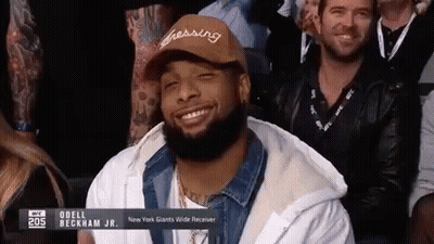 A Man With A Beard Wearing A Hat Is Smiling While Sitting In A Crowd . GIF