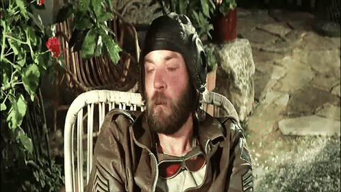 A Man With A Beard Wearing A Helmet And Sunglasses Is Sitting In A Chair GIF