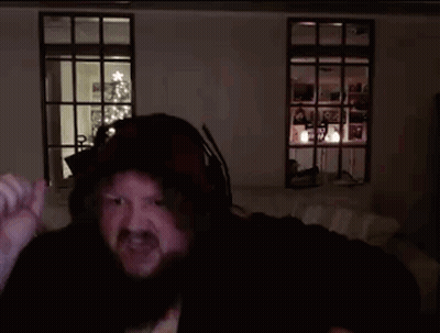 A Man With A Beard Wearing A Hooded Jacket Is Standing In A Dark Room With A Christmas Tree In The Background GIF