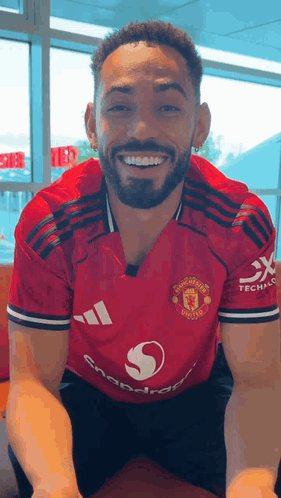 A Man With A Beard Wearing A Red Adidas Jersey Is Smiling While Sitting Down . GIF